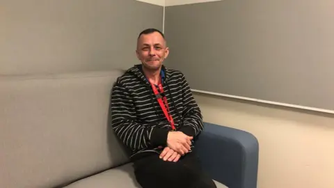 Vikki Irwin/BBC Dr. Granville Sutton sits on a sofa in an interview room at Martlesham Police's investigation centre. He is sitting on a comfortable couch, wearing a black and white striped sweater and dark pants.  