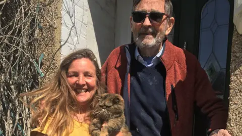 A woman in a yellow top and a man in a red jacket and sunglasses smile in the sun outside a front door. The woman is holding a cat, which looks grumpy.