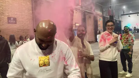 Amanda Harper A man in a white jumper stands in a concrete warehouse. A man behind him throws pink powder on him while a woman nearby watches on. 