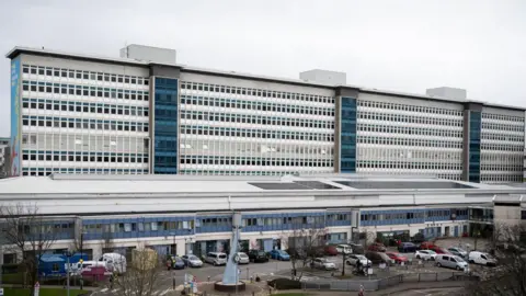 The outside of a large hospital, which is tall, blue and white with many windows, and car parked in front of it.