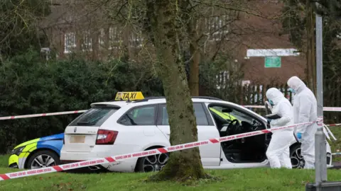 Pacemaker A white estate car with a taxi sign sits in a car park. There is a police cordon tied to a street sign. A marked police car is parked behind the taxi. There are a variety of trees and patches of grass at the perimeter. Two forensic officers in white body suits examine the taxi.
