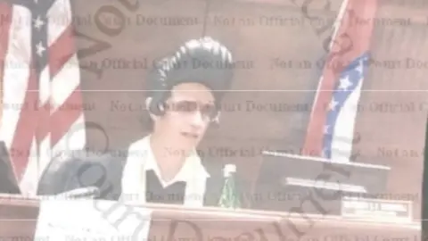 The State of Missouri's Commission on Retirement, Removal and Discipline of Judges An image of Judge Thornhill sitting at the court bench, taken from official court documents, showing him wearing an Elvis wig with the singer's signature black pompadour and sunglasses, as well as a traditional judge's robe with a white scarf around his neck. In front of him is a computer screen and a bottle of water. Behind him is a flag of the US and a flag of the State of Missouri. 