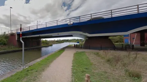 A view of the blue canal bridge from a footpath below it, on an overcast day.