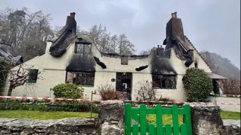 A burnt out cottage. The roof is gone, there is red and white tape in front of the building behind a green gate