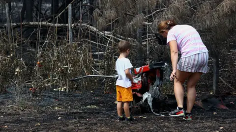 A woman and a toddler stand by charred ground and burned plants.