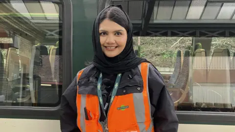 BBC, Leanne Rinne A woman wearing a black head scarf and orange high visibilty jacket standing on a platform in front of a train. 