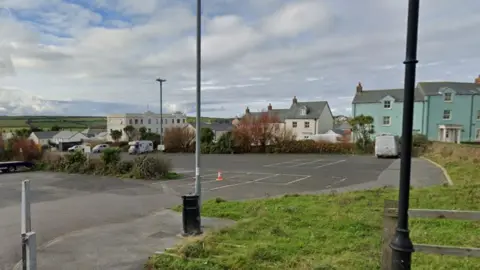The image shows a mostly empty tarmac car park with just a few vehicles scattered around, bordered by grass, lampposts and a single traffic cone near the marked bays. In the background there are pastel-coloured houses and low buildings, with open countryside stretching out under a cloudy sky.