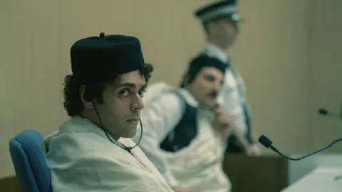 Carnival Film & Television Ardalan Esmaili, playing Megrahi, is in the foreground sitting in court listening through headphones. Mudar Abbara, playing Fhimah, is sitting in the background, and a police officer is standing in the distance. 