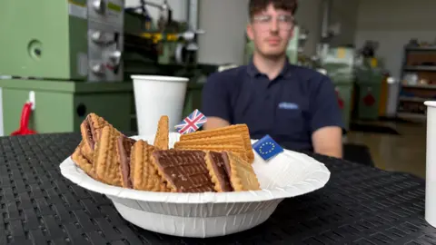Ben Schofield/BBC A paper bowl filled with rectangular biscuits that are chocolate-covered on one side. Also in the bowl are a mini Union Flag and a mini European Union Flag. The bowl is sitting on a black table. Out of focus in the background is a paper cup and further behind that a man is sitting down. Behind the man are large pieces of industrial equipment.