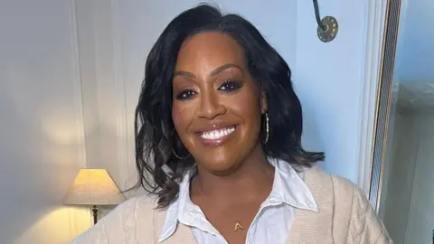 A woman smiles showing her teeth to the camera. She has shoulder-length black hair and wears a cream cardigan over a white blouse. Behind her is a white wall with the top of a white table lamp visible.