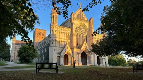 Simon Dedman/BBC St Albans Cathedral with the sun shining down on it. There are trees in the foreground and a bench, surrounded by grass, which faces the front of the cathedral