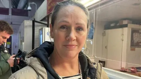 Louise Mortimer has blone hair in a bun and is wearing a fawn anorak,