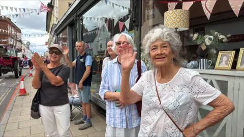 Richard Knights/BBC Men and women line the streets of a town and wave, clap and cheer. They are smiling.