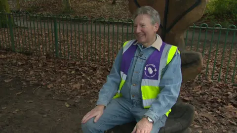 A man with grey hair sits on a sculpture within a park. He is wearing a light blue denim jacket and jeans as well as a yellow-and-purple hi-vis sleeveless jacket with the word 'volunteer' on it.