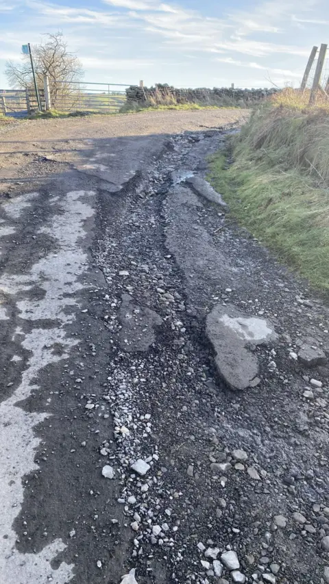 Jan Gilchrist A picture of a  country lane which has a long and narrow pothole and loose stones, a field gate and a footpath sign are visible in the background