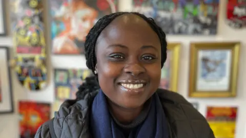 BBC Una mujer vestida con una chaqueta acolchada negra y una bufanda, sonriendo a la cámara. Detrás hay imágenes y fotografías desenfocadas.