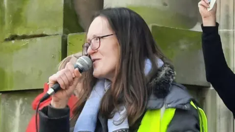 Emma Lochery wearing a hi-vis jacket and holding a mic. She is speaking at a campaign rally. 