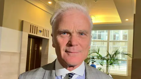 A close up image of the Conservative MP Sir Desmond Swayne looking straight to camera. He is standing in what looks like the lobby of an office building with a lift and potted plant in front of a window behind him. He is wearing a grey suit, blue and white pin striped shirt with a white collar, and black and white tie.