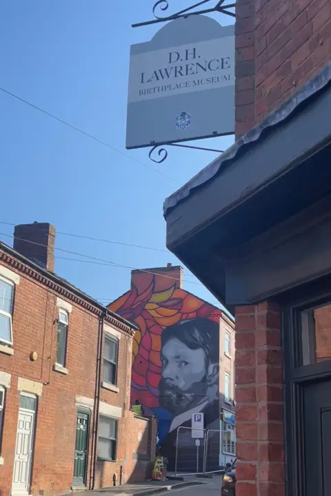 Nathan Bainbridge At the front of the photo is a sign outside the DH Lawrence Birthplace and Museum. In the background is the mural. 