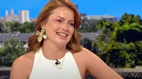 Tasha Ghouri, a young woman with blonde hair, wears a white top and smiles, sitting on the BBC Breakfast sofa 