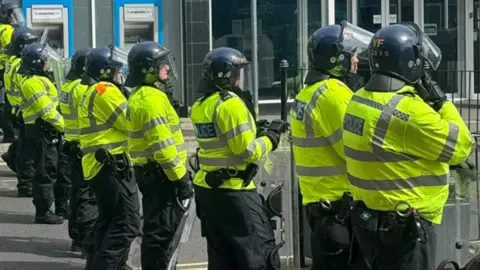 BBC A line of police officers in riot gear, wearing helmets and holding shields
