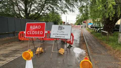 Tom Jackson/BBC A road leads to a level crossing which is in the background. There are pavements either sides of the road. In the foreground, there are different signs and cones across the road.