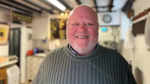 George Carden/BBC a bald man with a red face and white stubble is stood in a room with wooden beams, which could be the kitchen of a house or a business. He is smiling at the camera and wearing a grey ribbed jumper.