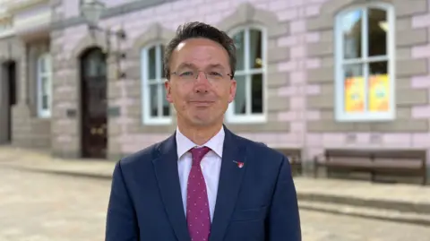 A person wearing a navy blue suit jacket, white shirt, and red patterned tie stands facing the camera in a paved courtyard. Behind the person is a pastel pink building with stone trim, arched windows, and a dark wooden doorway.