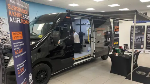 A black van with branding on it, parked inside a large room. There is a vertical banner on the left which says "surrender a knife, save a life today". there is a table set up outside the van under an awning.