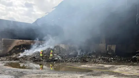 A large warehouse is seen gutted by fire. Outside the building two firefighters stand in front of piles of burned rubbish, wearing yellow hemlets and protective clothing. Smoke smoulders from the pile of rubbish and spreads into the air, covering much of the photo. There is a wide puddle of water showing the firefighters' reflections.