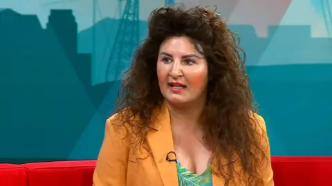 Natalie Younes is also in the red Politics North studio. She has brown, curly hair and is wearing a yellow blazer.