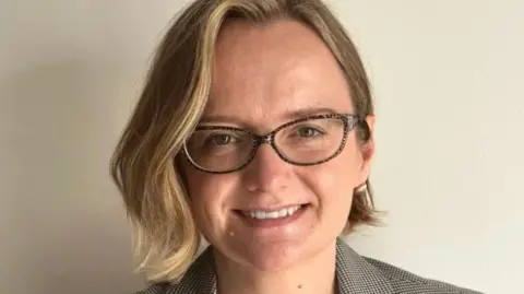 A woman with short blonde hair and glasses smiles at the camera. She is standing in front of a white wall. 