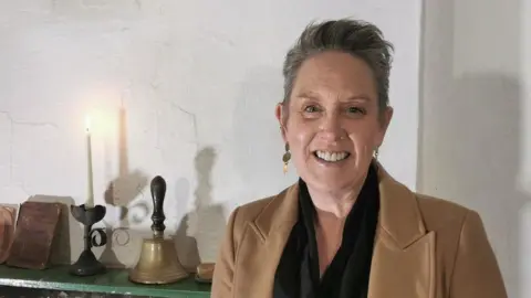 A lady with a short grey quiff stands in front of a whitewash wall, smiling. She wears a tan woollen coat with lapels, and a black scarf.