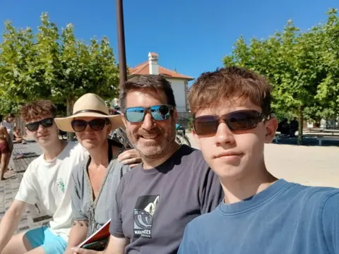 David Morton A family picture - two teenage sons are sat either side of their parents with blue sky and trees in the background. All four family members are wearing T-shirts and sunglasses.
