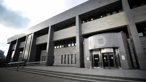 PA Media A general view of Glasgow Sheriff Court, a grey building with a grey exterior and a entrance with a revolving door which has a crest above it.