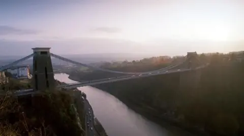 A panoramic view of the Clifton Suspension Bridge in the early morning light. The Bridge has two huge towers at each end. The River Avon is in visible in the gorge below.