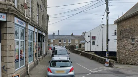 A general view of the bridge. Cars are parked on the left hand side of the road which leads to the bridge. Buildings are built up around it. 