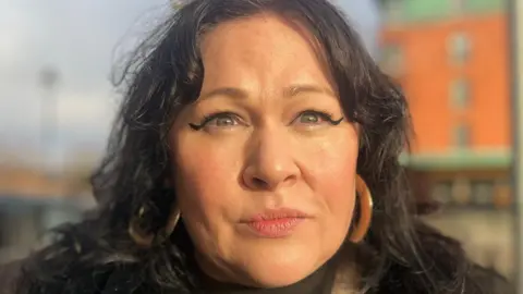 BBC Sara Morrison has long dark hair parted in the middle and is wearing large hoop ear-rings