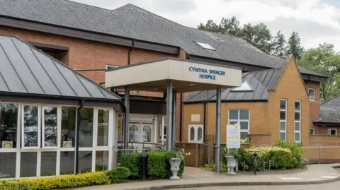 Cynthia Spencer Hospice The outside of Cynthia Spencer Hospice, with a canopy, and plant pots and foliage. The building is made of brick, with a large roof and glasses building to the left.