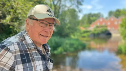 Owen Sennitt/BBC A man wearing a baseball cap and glasses and shirt looks towards the camera, with a river and mill blurred in the background.