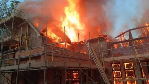 HIWFRS Flames and smoke inside and from roof of house partially built - it is surrounded by scaffolding and is built with large grey blocks/bricks.