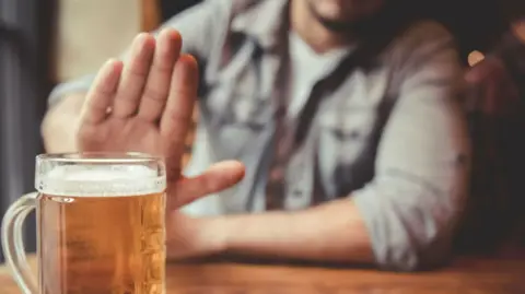 A close up of a pint of beer. A man wearing a denim jacket is blurry in background holding his hand up seemingly "rejecting" the beer.