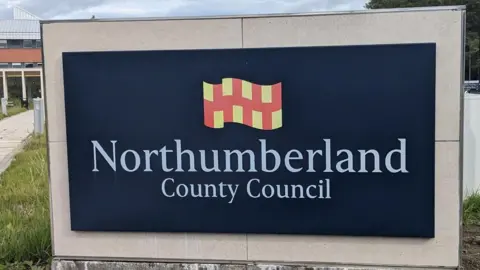 A black sign with white writing which reads 'Northumberland County Council' which sits underneath a red and yellow flag. 