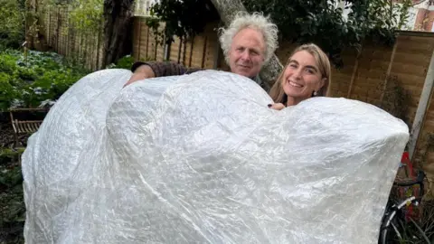 A picture of Charlie Mackesy and Rachael de Caux, who are stood behind an elephant that is covered by bubblewrap or some sort of material.