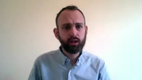 A man with a beard wearing a light blue shirt looks into the camera.