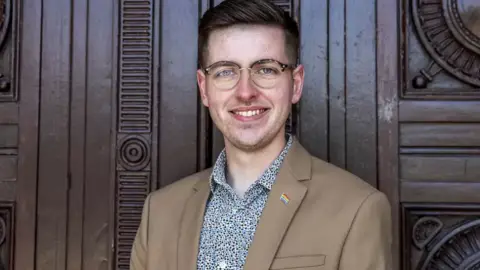 Liverpool City Council Harry Doyle, who has dark blonde hair swept to the side and round framed glasses, and is wearing a tan coloured blazer over an open-collared shirt with a multi-coloured dot pattern, smiles at the camera standing against a dark wooden wall. 