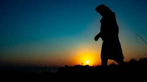A single lady walks in the hills of Lesvos island to avoid the police. Silhouettes of walking refugees and migrants on the hills early in the morning during the dawn and the sunrise over the Aegean sea and Turkey