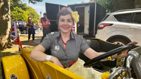 A woman with dark hair, a headscarf and large yellow flower smiles as she sits in black and yellow car. She is wearing a black check dress with a red necklace and red belt.
