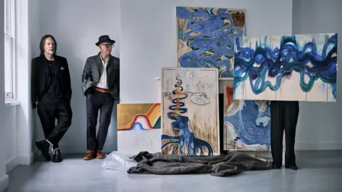 Julian Broad Thom Yorke and Stanley Donwood are stood leaning against the walls of an alcove in a large white room. Donwood is wearing a trilby hat and glasses. Yorke has long hair and a white short beard. Next to them is a collection of five abstract paintings.