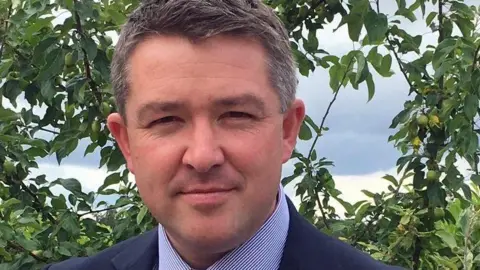 A man with short dark grey hair, wearing a dark blue suit and a blue pinstripe shirt, looks at the camera. He is standing in front of a fruit tree of some kind.